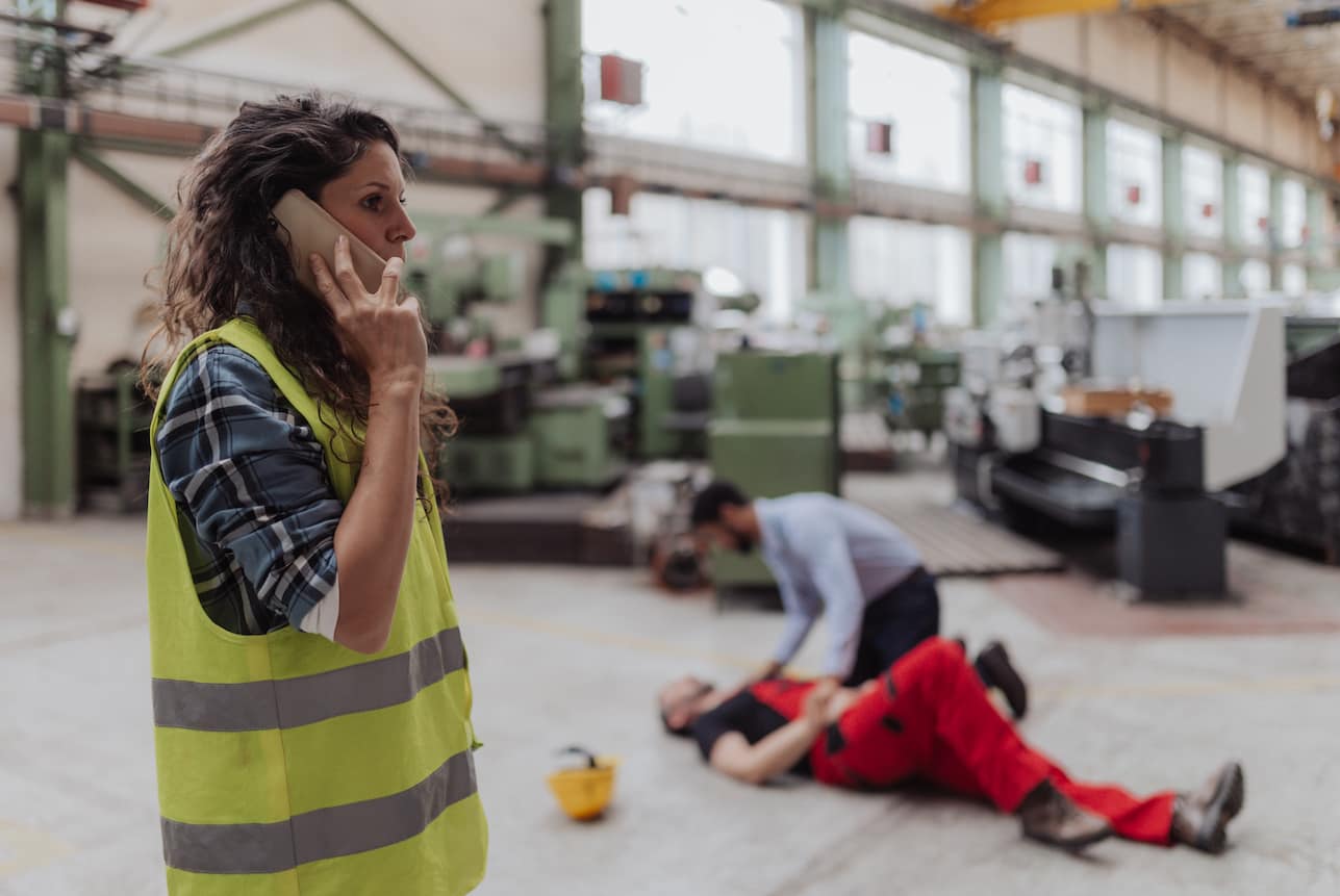 Imagen de un accidente laboral en el que se observa una persona tirada en el suelo siendo asistida por un compañero de trabajo mientras que otra persona llamada a emergencias. 